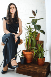 Portrait of jazz vocalist Issey Chivers seated beside potted plants against a white brick wall.